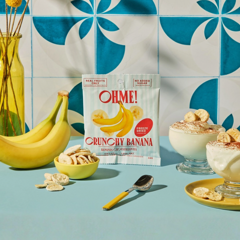 Packaging of OHME! Crunchy Banana with bananas, nuts, and dessert on a blue surface with a tiled wall background.