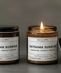 Two candles labeled 'Satsuma Sunrise' on a gray surface with a blurred background