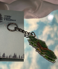 Keychain with nature-themed design on a card with 'Tide and Timber' branding, against a blurred natural background.