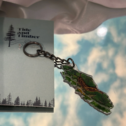 Keychain with nature-themed design on a card with 'Tide and Timber' branding, against a blurred natural background.