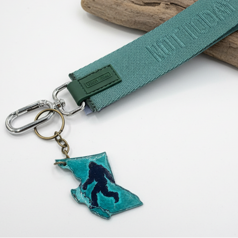 Keychain with teal ribbon, keyring, and Bigfoot silhouette on a wooden surface