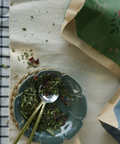 Tea leaves in a bowl with a spoon on a checkered cloth, next to tea packages.