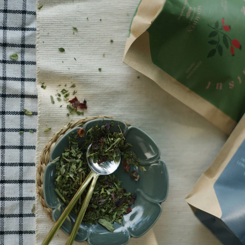Tea leaves in a bowl with a spoon on a checkered cloth, next to tea packages.
