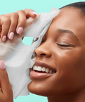 Woman applying a white sheet mask to her face against a teal background