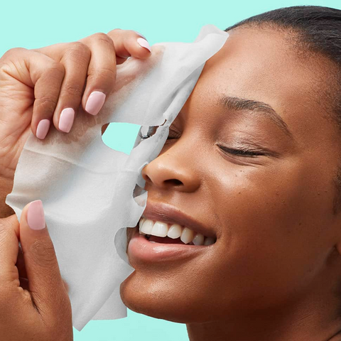 Woman applying a white sheet mask to her face against a teal background