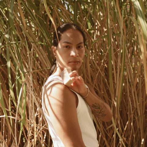 Person standing among tall grass wearing a white sleeveless shirt and brown pants.