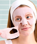 Woman applying a pink face mask with a white headband against a light blue tiled wall.
