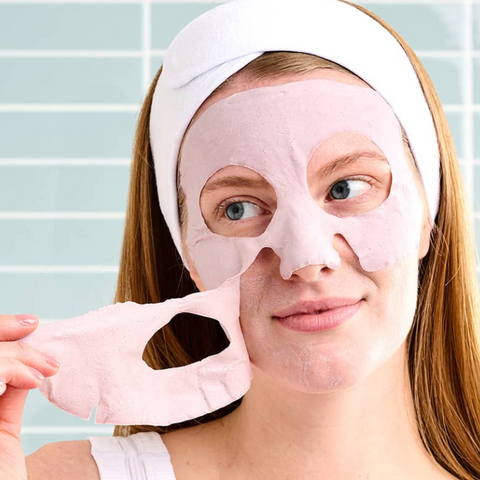 Woman applying a pink face mask with a white headband against a light blue tiled wall.