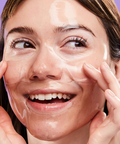 Woman applying a skincare product on her face with a purple background