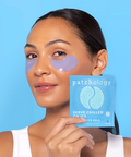 Woman wearing eye patches holding a box of patchology eye gels against a blue background