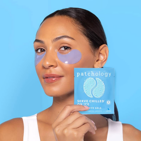 Woman wearing eye patches holding a box of patchology eye gels against a blue background