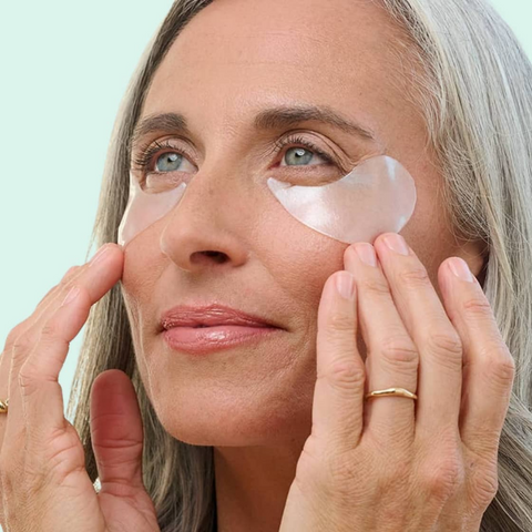 Woman applying pink eye patches to her eyes with a light blue background