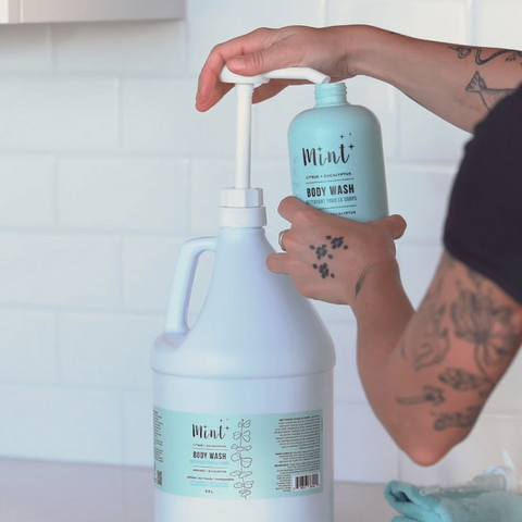Person pouring Mint body wash from a smaller bottle into a larger container.