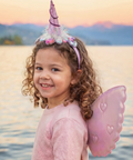 A young girl with curly brown hair wearing a pink unicorn costume accessory set, featuring a pink horn headband with a feathered top and translucent pink wings, posing outdoors in front of a lake at sunset.