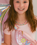 A young girl with long light brown hair smiles while wearing a shiny pink fanny pack with colorful 'OK' letter patches. She is wearing a light pink t-shirt and a heart-patterned necklace, and a large iridescent balloon is visible on the left side of the frame.