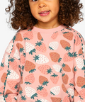 A young child with curly brown hair wearing a pink crewneck sweatshirt with a strawberry pattern, photographed against a light gray studio background.