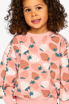 A young child with curly brown hair wearing a pink crewneck sweatshirt with a strawberry pattern, photographed against a light gray studio background.