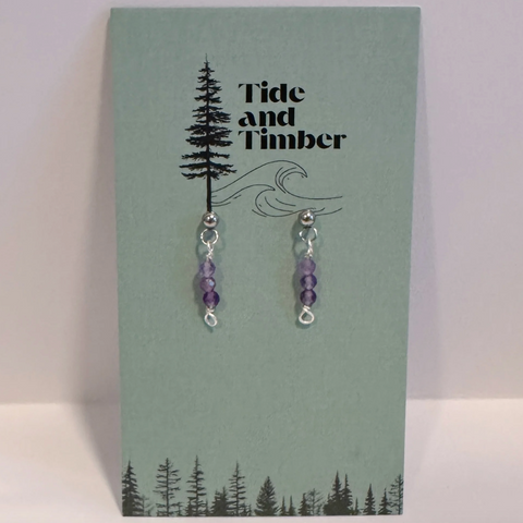 Pair of purple beaded earrings on a Tide and Timber branded card with tree and wave design.