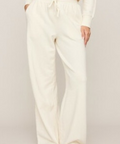 A person wearing cream-colored wide-leg sweatpants with a drawstring waist, paired with a matching cream sweatshirt and tan slippers, shown against a plain light beige studio background.