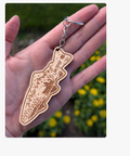 A hand holding a wooden arrowhead-shaped keychain with a silver metal ring, set against a blurred background of green foliage and yellow flowers.