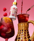 Bottle of Sangria Directa being poured into a glass with fruit and a pitcher on a pink background