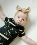 Baby wearing a black outfit with gold patterns and a large bow on a white background