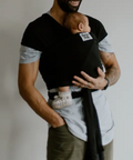 Man holding a baby in a black baby carrier against a plain background