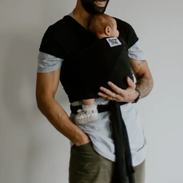 Man holding a baby in a black baby carrier against a plain background