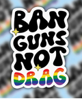 Sticker with 'Ban Guns Not Drag' text and rainbow colors on a blurred background