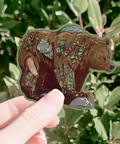 Hand holding a bear sticker with nature-themed design against a green leafy background