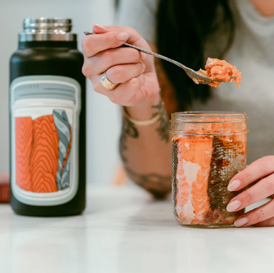 Person holding a jar of salmon and a bottle with a label, sitting at a table.