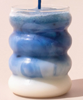 A textured, ribbed glass tumbler with a wavy, stacked design, filled with a blue and white marbled beverage and topped with a blue straw, set against a solid pale pink background.