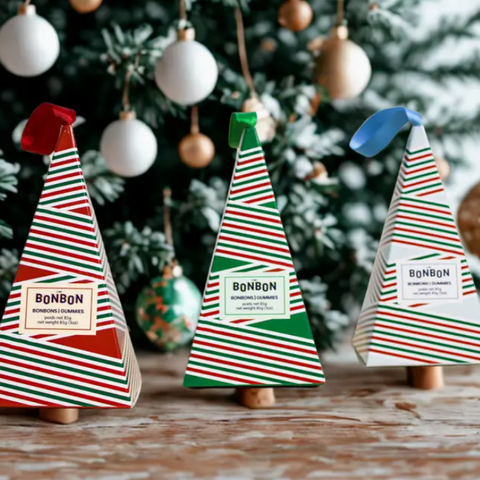 Three decorative Christmas tree-shaped boxes with striped patterns and 'Bonbon' labels in front of a Christmas tree.