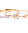 A pair of gold bangle bracelets, one adorned with a row of iridescent clear crystals and the other decorated with two matching crystal flower charms, set against a plain white background.