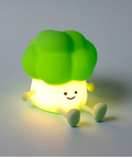 A cute broccoli-shaped night light glowing with a warm yellow light, featuring a bright green floret top and a smiling face on a soft gray background.