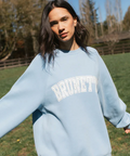 Woman wearing a light blue sweatshirt with 'BRUNETTE' text, standing outdoors in a grassy area.