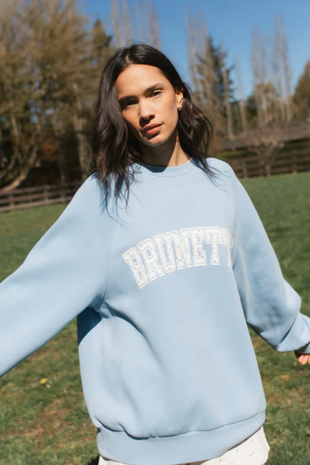 Woman wearing a light blue sweatshirt with 'BRUNETTE' text, standing outdoors in a grassy area.