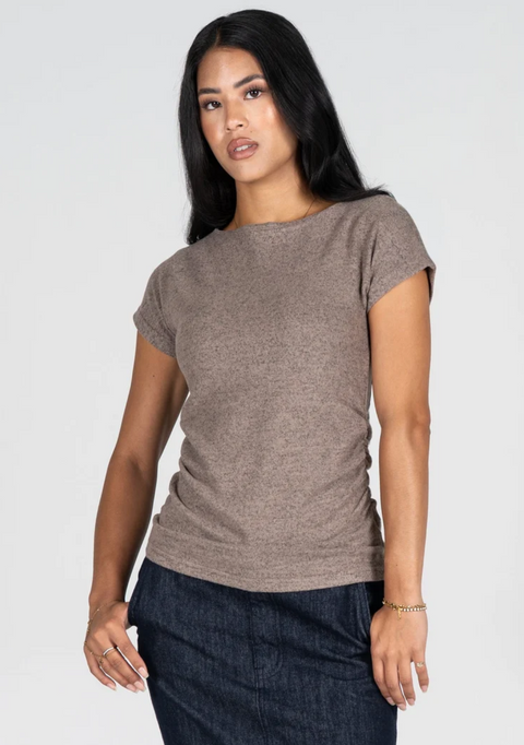 A woman modeling a short-sleeve crew-neck knit top in taupe brown, paired with dark denim pants, against a plain light gray studio background.