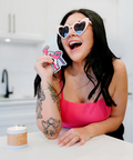 Woman wearing heart-shaped sunglasses holding a pink bow card in a kitchen.