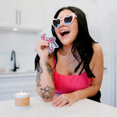 Woman wearing heart-shaped sunglasses holding a pink bow card in a kitchen.