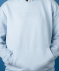 A person wearing a light blue pullover hoodie with the words 'CAN YOU TELL I'M ANXIOUS?' printed in tonal, slightly darker blue text on the chest, set against a solid blue background.
