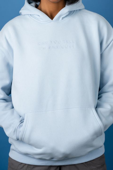 A person wearing a light blue pullover hoodie with the words 'CAN YOU TELL I'M ANXIOUS?' printed in tonal, slightly darker blue text on the chest, set against a solid blue background.