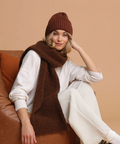 Woman wearing a brown knit hat and scarf sitting on a brown couch against a beige background