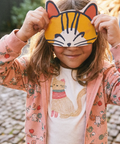A young child wearing a pink floral zip-up jacket over a white t-shirt with a cat graphic, holding a yellow cat-face mask in front of their eyes. The background shows a blurred outdoor cobblestone path.