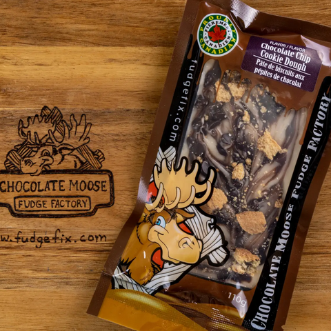 A 10g bag of Chocolate Moose Fudge Factory Chocolate Chip Cookie Dough fudge, placed on a wooden surface next to a laser-engraved logo of the same brand.
