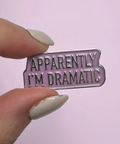 Enamel pin with 'Apparently I'm Dramatic' text held by a hand against a light purple background