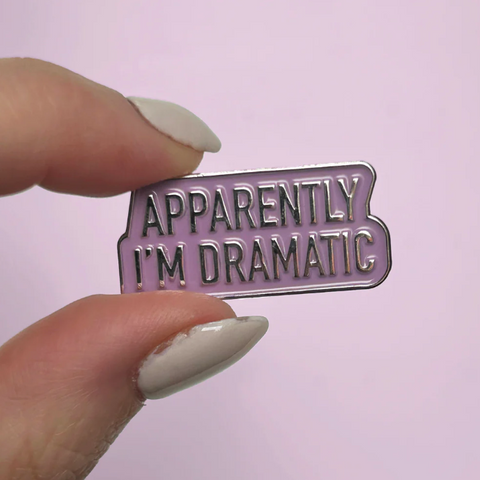 Enamel pin with 'Apparently I'm Dramatic' text held by a hand against a light purple background