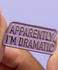 Enamel pin with 'Apparently I'm Dramatic' text held between fingers against a purple background