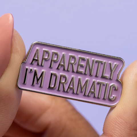 Enamel pin with 'Apparently I'm Dramatic' text held between fingers against a purple background