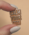Enamel pin with 'CALM YOUR TITS' text held between fingers against a beige background